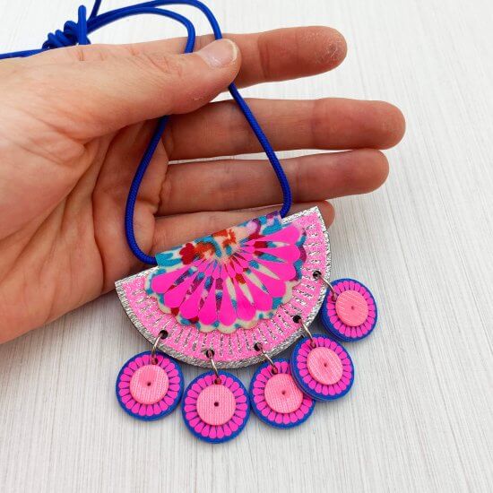A pink, silver and blue jangling charm necklace is held in a white hand against an off white background