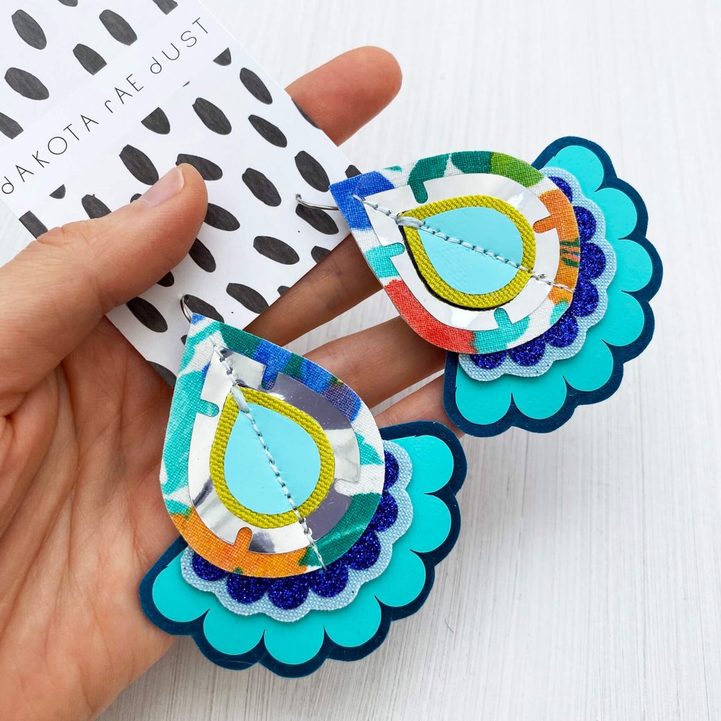 A pair of turquoise, silver and blue vintage fabric teardrop earrings mounted on a dakota rae dust branded card, held in a woman's hand