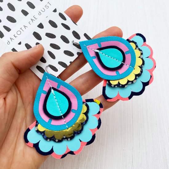 A pair of teardrop shaped, oversize turquoise earrings mounted on a black and white patterned, dakota rae dust branded card are held in a white open hand.
