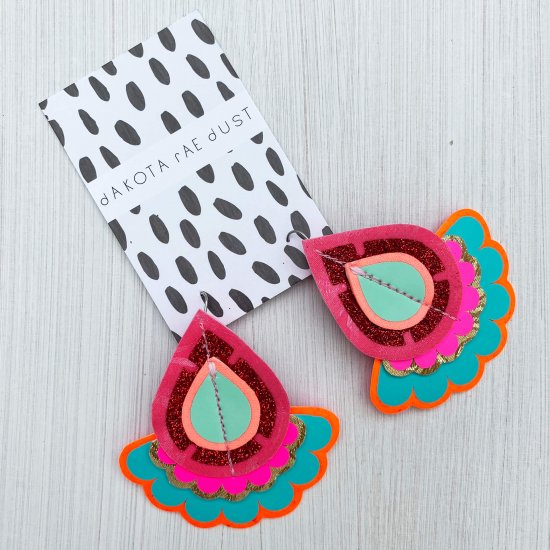 A pair of pink and turquoise colourful oversize earrings mounted on a black and white patterned, dakota rae dust branded card are lying on an off white background
