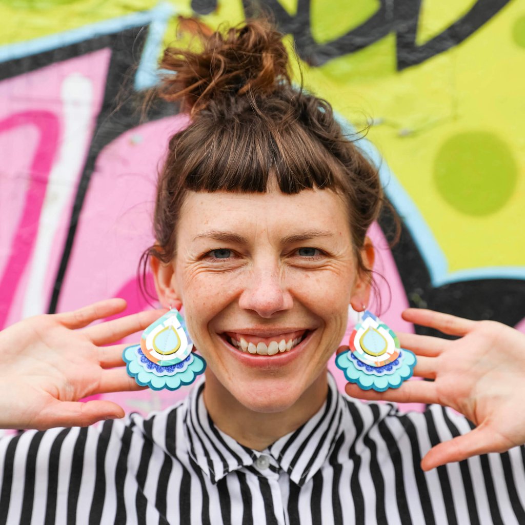 A white woman with a short fringe is facing the camera smiling. She is wearing a large pair of colourful earrings and holding a hand behind each ear to keep them facing towards the camera and draw attention to them. She is wearing a black and white stripey shirt and behind her the wall is spray painted in lime and pink patterns