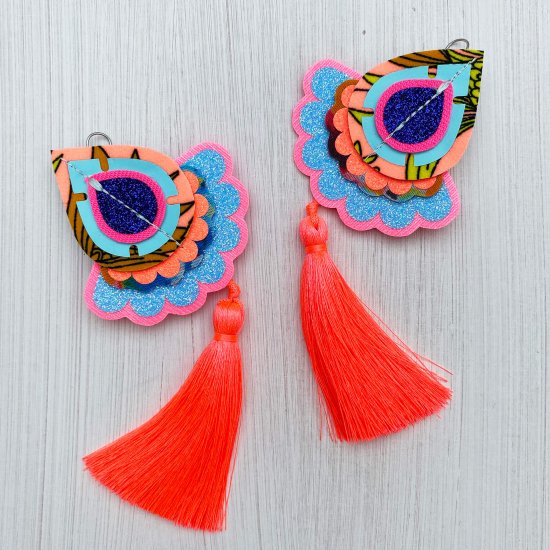 A pair of decorative teardrop shaped, neon coral oversize earrings with silky tassels are lying on an off white background. tassel earrings