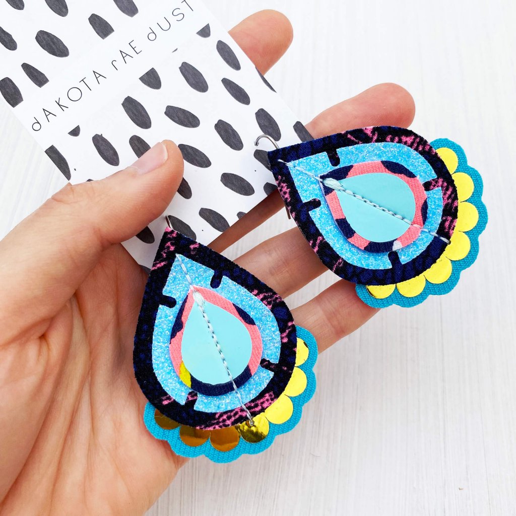 A pair of colourful recycled fabric earrings in baby blue, navy and gold held in a woman's open hand