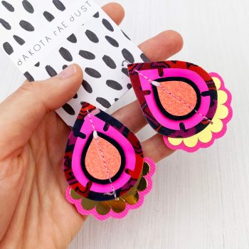 A pair of neon pink, coral and gold teardrop shaped, jazzy plectrum earrings held in a woman's open hand