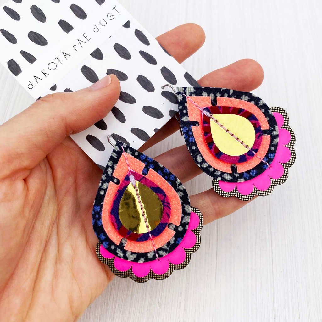 A pair of glittery coral, neon pink, navy and gold classic teardrop earrings held in a woman's open hand