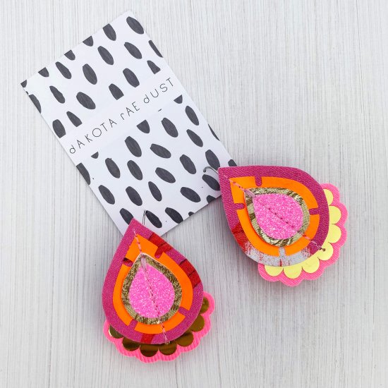 A pair of pink, orange and gold teardrop shaped, jazzy plectrum earrings mounted on a black and white patterned, dakota rae dust branded card.