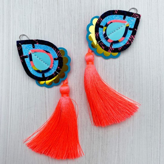 A pair of turquoise, neon coral and gold teardrop tassel earrings lying on a plain off white background.