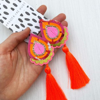A pair of bright pink and orange tassel earrings mounted on a black and white patterned, dakota rae dust branded card, held in a woman's hand.