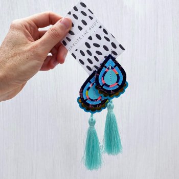 A pair of bubblegum blue glitter and turquoise tassel earrings mounted on a black and white patterned, dakota rae dust branded card, held in a woman's hand.