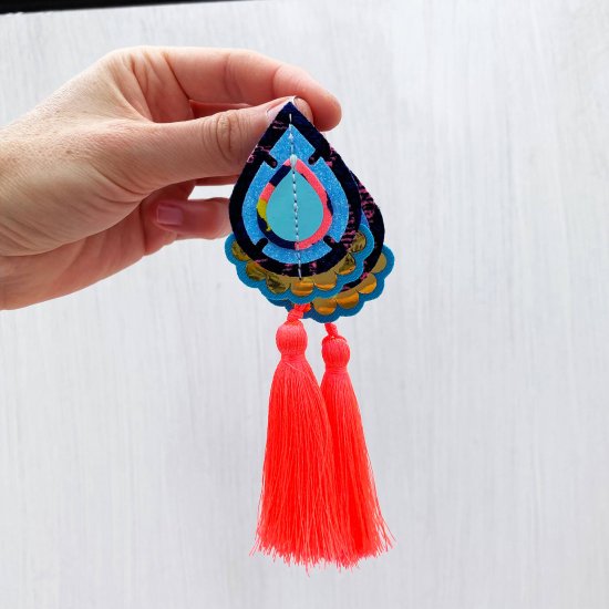 A pair of turquoise, neon coral and gold teardrop tassel earrings held in a white hand in front of a plain off white background.