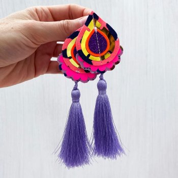 A pair of neon coral, gold and lilac tassel earrings are held in a woman's hand.