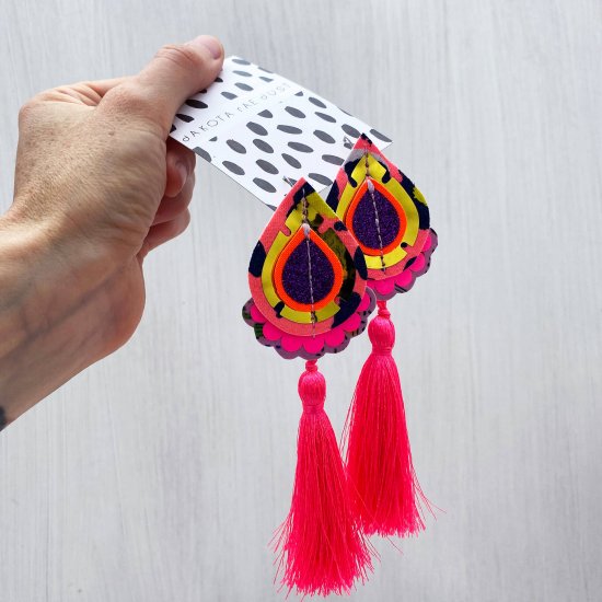 A pair of neon coral, gold and pink tassel earrings mounted on a black and white patterned, dakota rae dust branded card, held in a woman's hand.