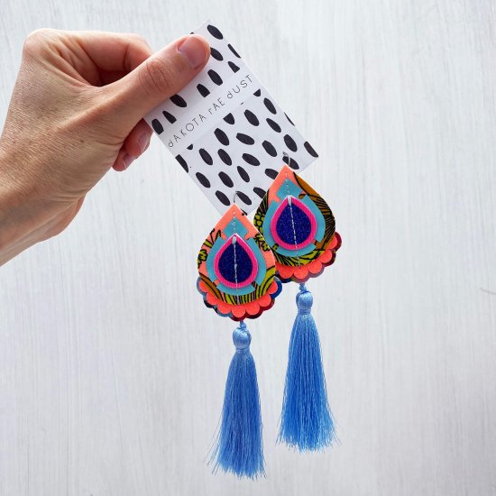 A pair of soft blue and peach teardrop tassel earrings mounted on a black and white patterned card, held by a woman's hand in front of a plain off white background.
