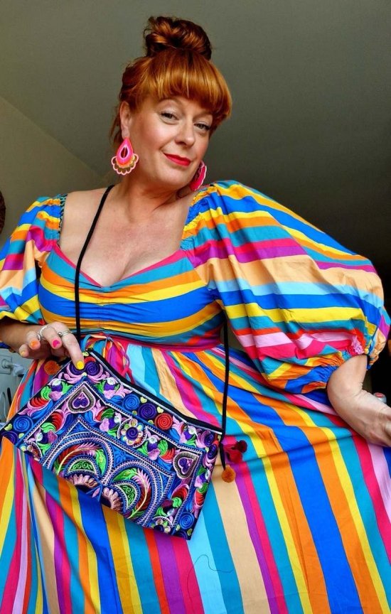 A woman with coppery coloured hair piled in a bun and a shiny fringe is posing in a colourful rainbow striped dress, embroidered shoulder bag and large statement earrings