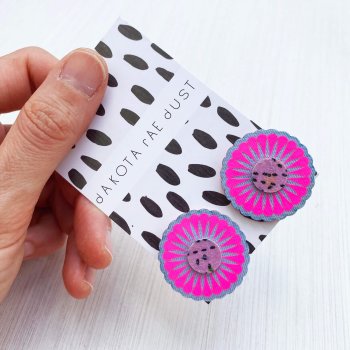 a set of oversize neon pink studs mounted on a black and white patterned, dakota rae dust branded card are held between a woman's thumb and forefinger against an off white background