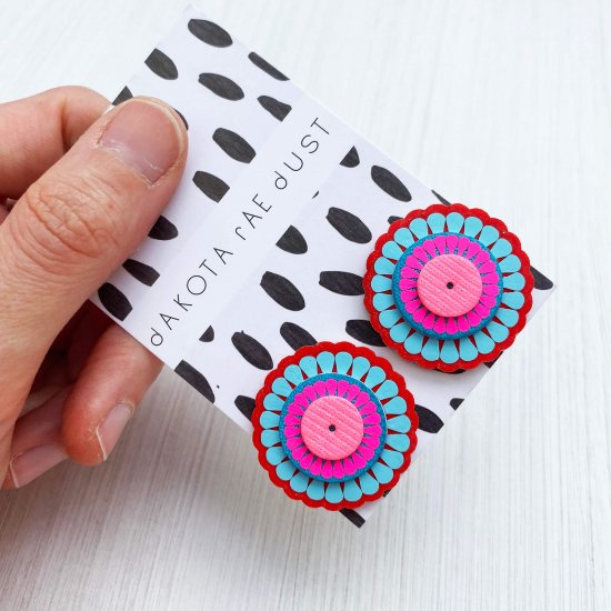 A colourful pair of oversize, circular ornate studs mounted on a black and white patterned, dakota rae dust branded card are held up to the camera by a white hand. Only the thumb and fingers are visible.