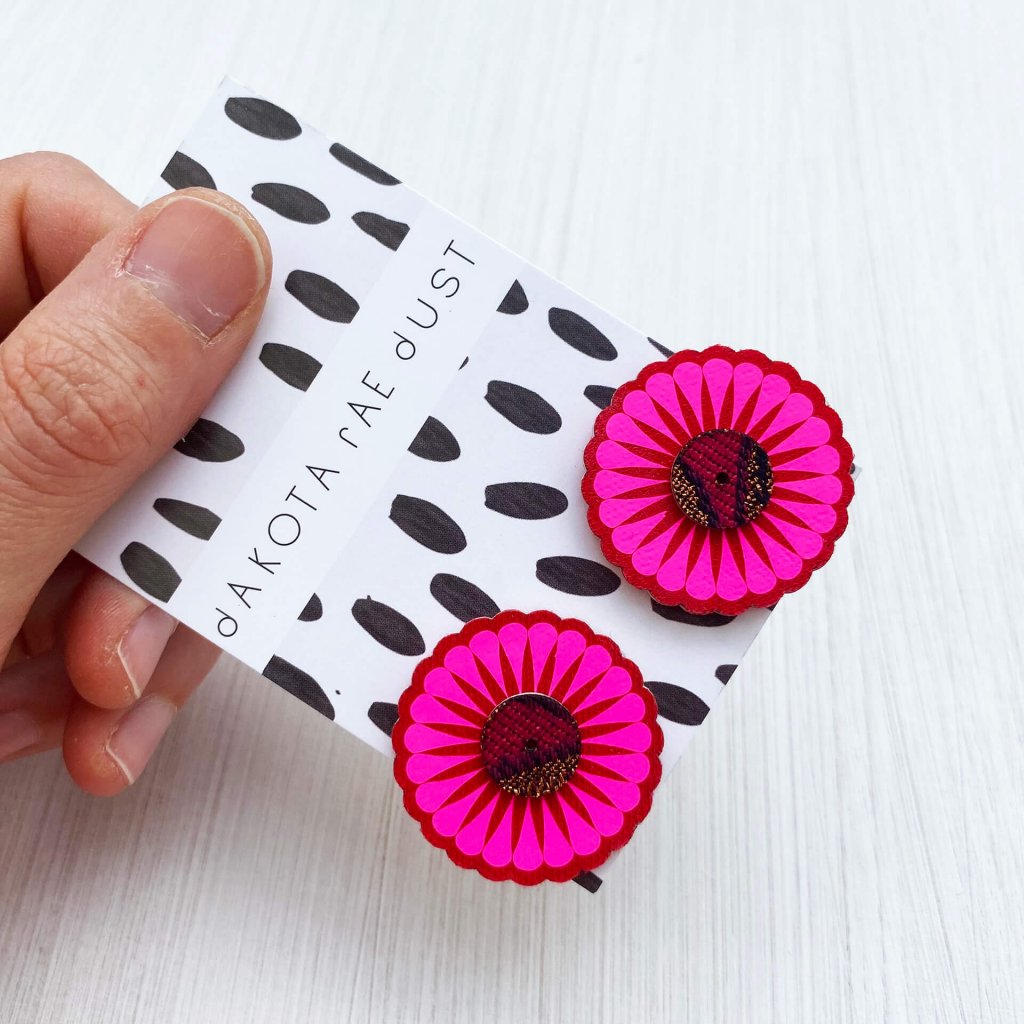 a set of red and fluorescent pink oversize studs mounted on a black and white patterned, dakota rae dust branded card are held between a woman's thumb and forefinger against an off white background