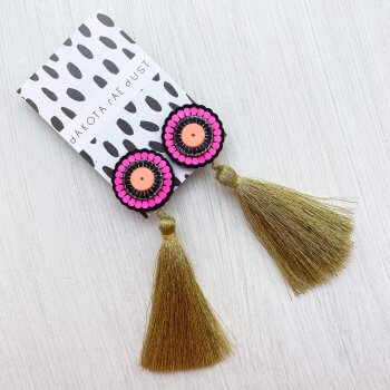 A pair of oversize tassel studs in black, neon pink and gold, mounted on a black and white patterned dakota rae dust branded card are seen on an off white textured background