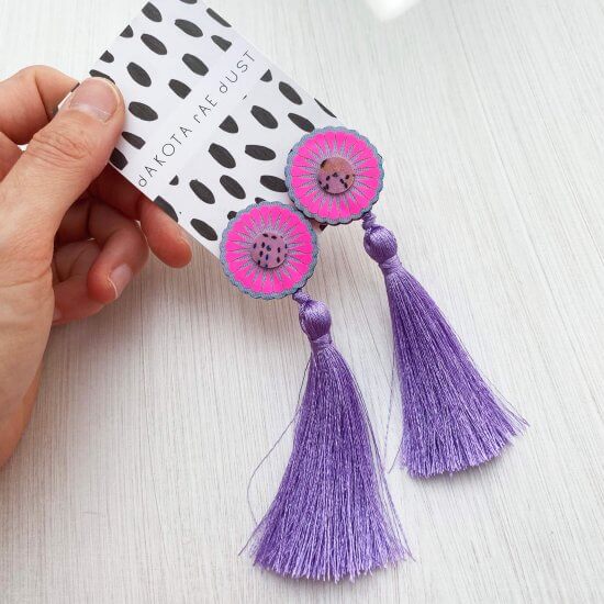 a pair of oversize tassel studs backed on a black and white patterned, dakota rae dust branded card are held by a woman's hand, just visible in the top left of the photo