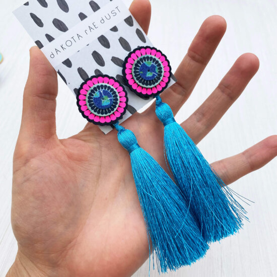 A pair of oversize tassel studs in turquoise, navy and neon pink are held in a woman's open hand.