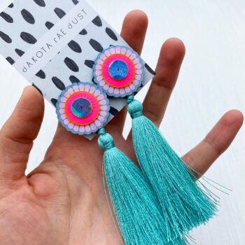A pair of oversize tassel studs in green, pale pink, neon pink and blue, mounted on a black and white patterned dakota rae dust branded card are held in an open hand