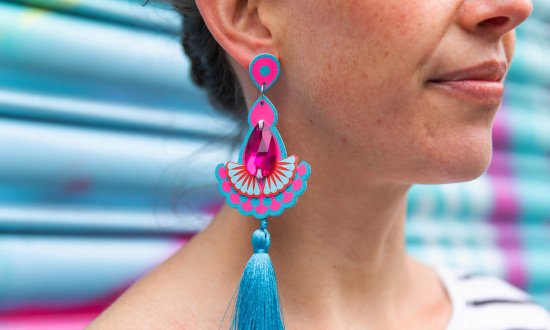 A close up of a woman's lower face and neckl focusing on her colourful statement tassel earrings. Behind her is a turquoise and pink spray painted metal shutter