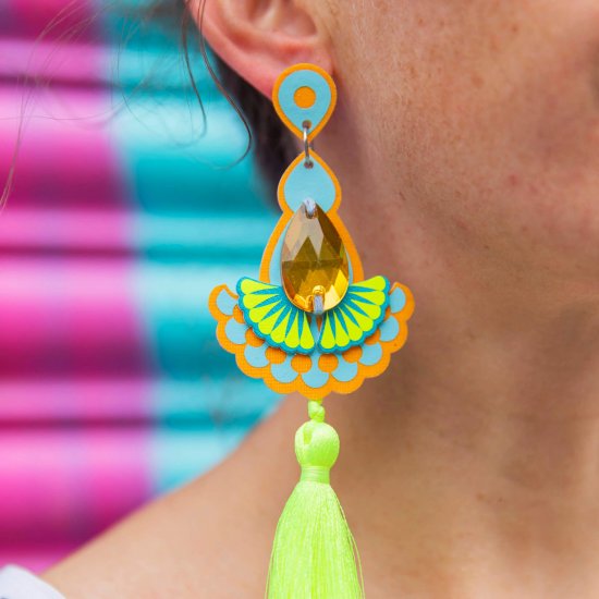 A close up of a woman's ear and neck focusing on the colourful tassel earrings in yellow and blue that she is wearing. The background of the image is a bright turquoise and pink metal shutter