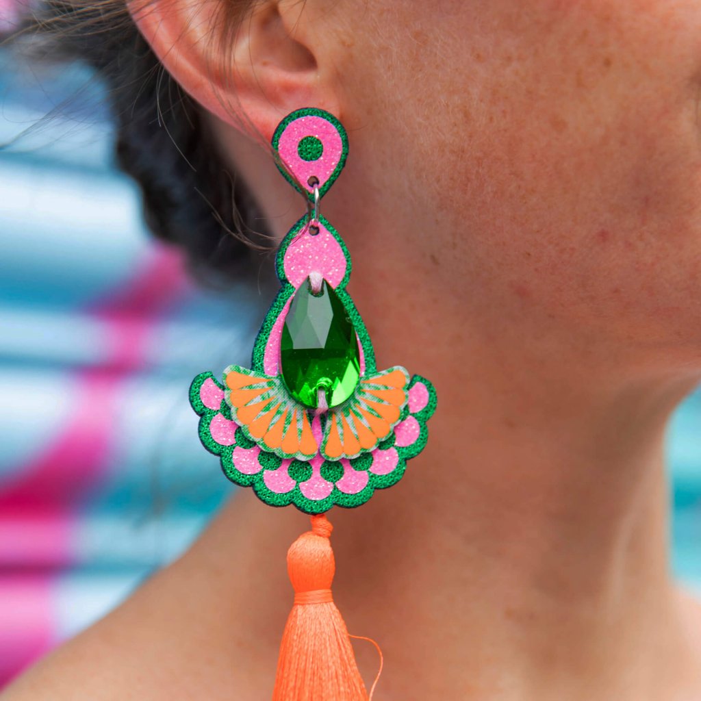 A close up of a woman's ear and neck focusing on the luxury tassel earrings in emerald green, pink and orange that she is wearing. The background of the image is a bright turquoise metal shutter