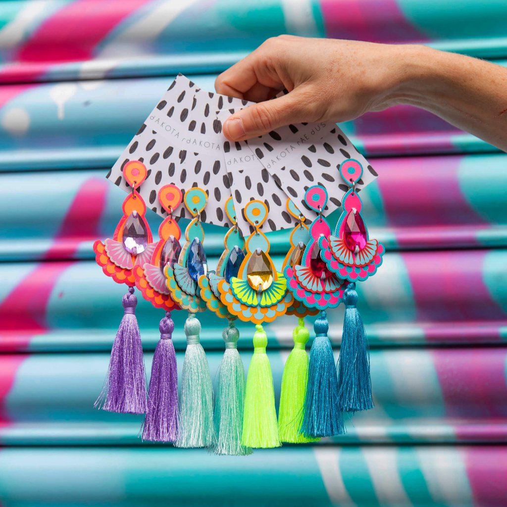 A hand holds a bunch of colourful statement tassel earrings in front of some colourful street art in turquoise and pink