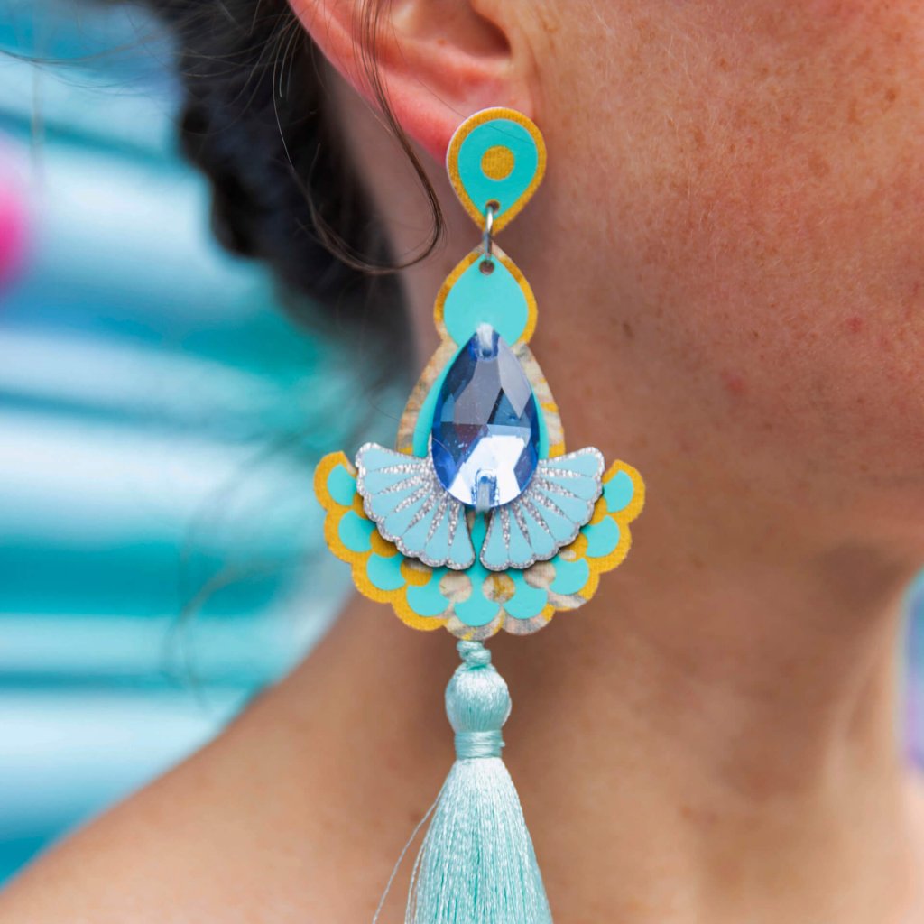 A close up of a woman's neck and ear focusing on her yellow and pale turquoise luxury tassel earrings adorned with mid blue teardrop shaped jewels