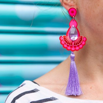 A close up of a woman's ear and neck focusing on the luxury tassel earrings in red, pink and lilac that she is wearing. The background of the image is a bright turquoise metal shutter
