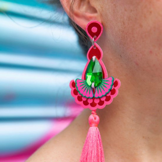A close up of a woman's neck and ear focusing on her pink earrings with tassels. The earrings have large teardrop shaped emerald green jewels and silky neon pink tassels