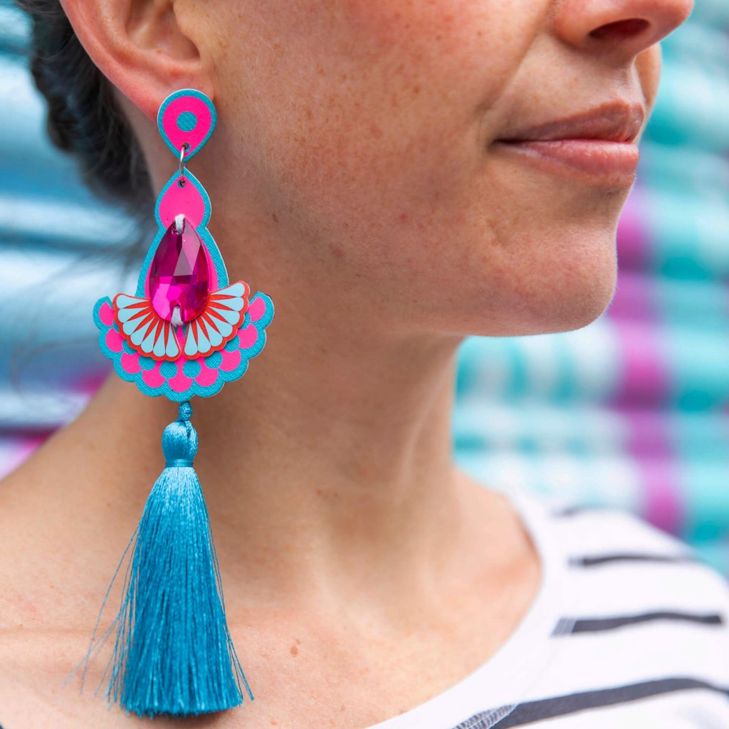 A close up of the lower half of a woman's face and neck focusing on the bright blue and neon pink colourful tassel earrings she is wearing. She is wearing a black and white stripey T-shirt which is just visible on her shoulder and is standing in front of a colourful turquoise and pink spray painted metal shutter