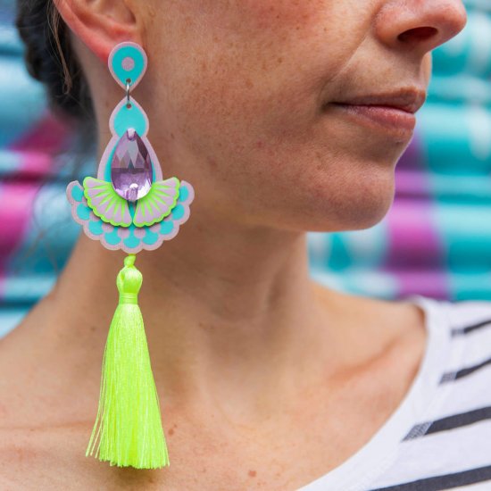 A close up of the lower half of a woman's face and neck focusing on the luxury neon tassel earrings she is wearing. She is wearing a black and white stripey T-shirt which is just visible on her shoulder and is standing in front of a colourful turquoise and pink spray painted metal shutter