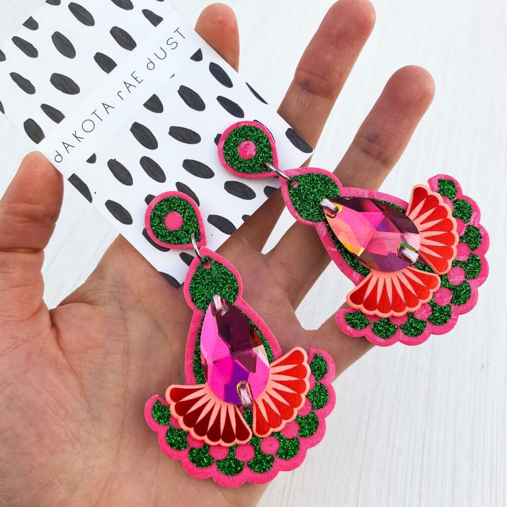 A pair of pink, red and emerald green glittery earrings held in a woman's open hand