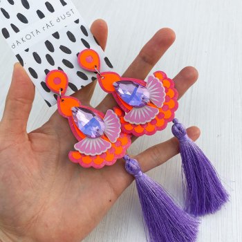 A pair of luxury tassel earrings held in an open hand, against an off white background. The orange and pink earrings have lilac tassels.