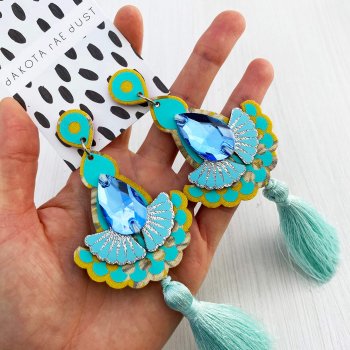 A pair of yellow and pale turquoise luxury tassel earrings adorned with mid blue teardrop shaped jewels are held in a open hand against an off white background