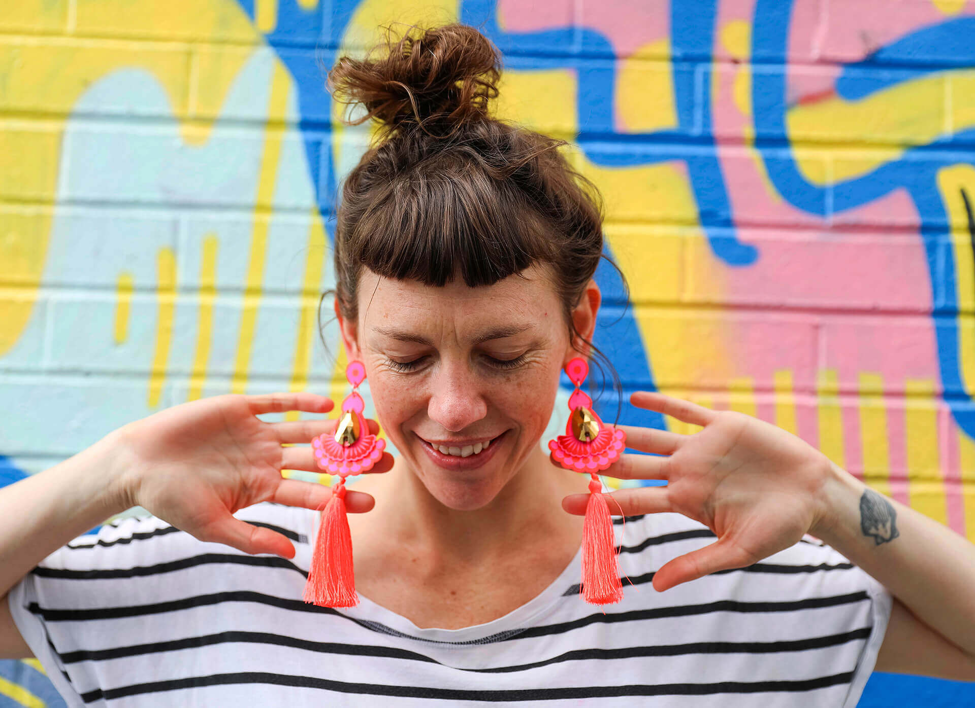 A woman with a short brown fringe wearing a black and white stripey t-shirt and large neon orange and pink statement tassel earrings is standing in front of a colourful graffiti covered wall. Her head is bowed and she is holding an open hand behind each ear to draw attention to her earrings