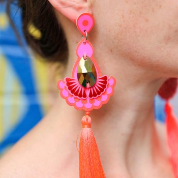 A close up of a woman's ear and neck, focusing on her fluorescent pink and orange statement tassel earrings which feature teardrop shaped gold jewels