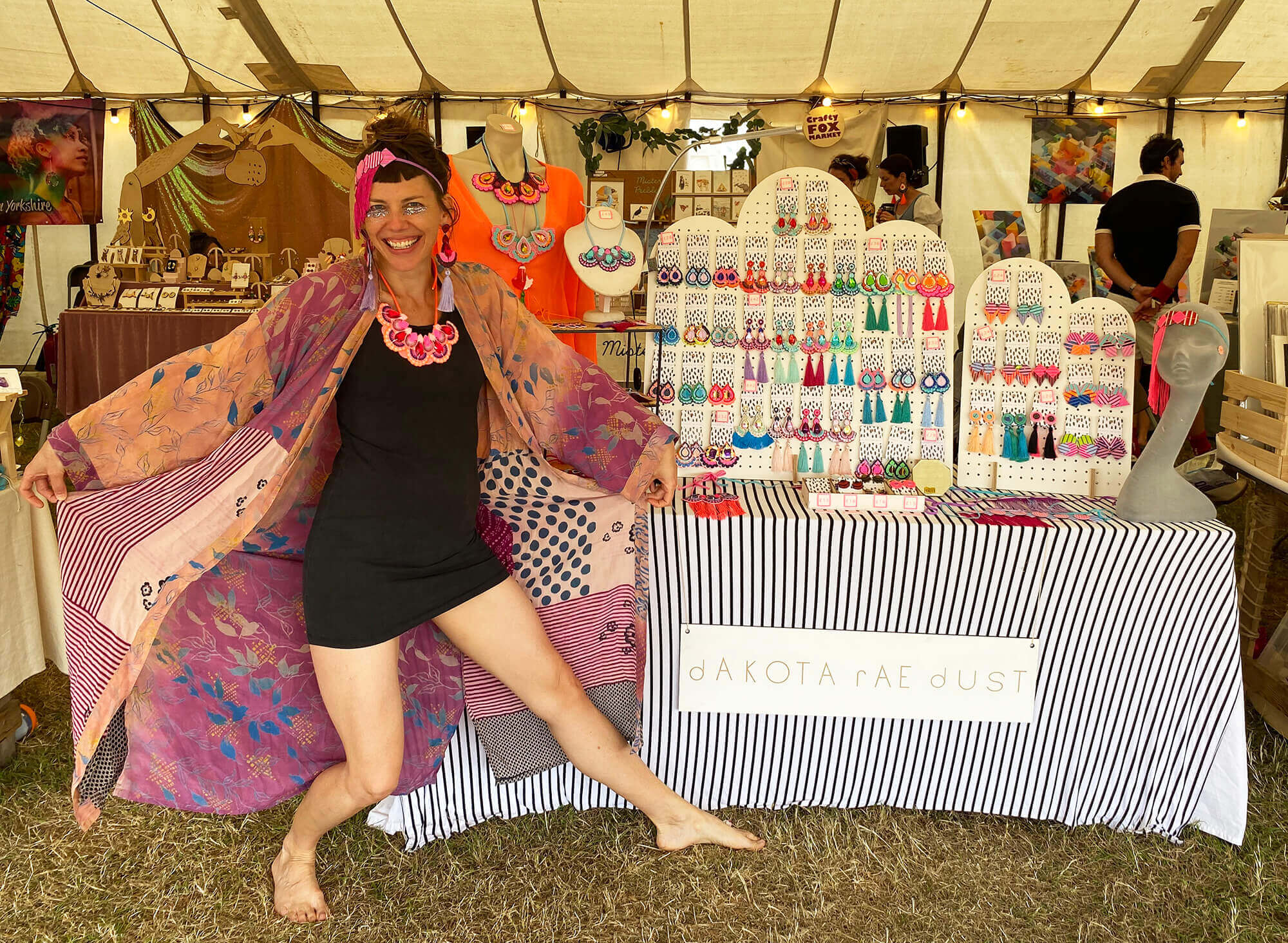 A woman wearing a fringed headdress, colourful bib necklace and patterned purple gown is posing in front of a colourful market stall. The white pegboards on the table are displaying rows of colourful festival jewellery, tassel earrings and bib necklaces.