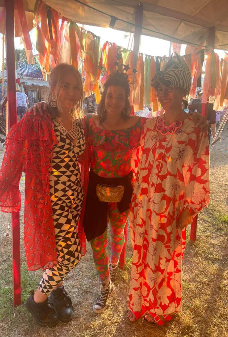 A warmly lit photo of three women dressed in brightly patterned outfits inside a marquee with colourful tassel bunting. The women have their arms around each other and are smiling