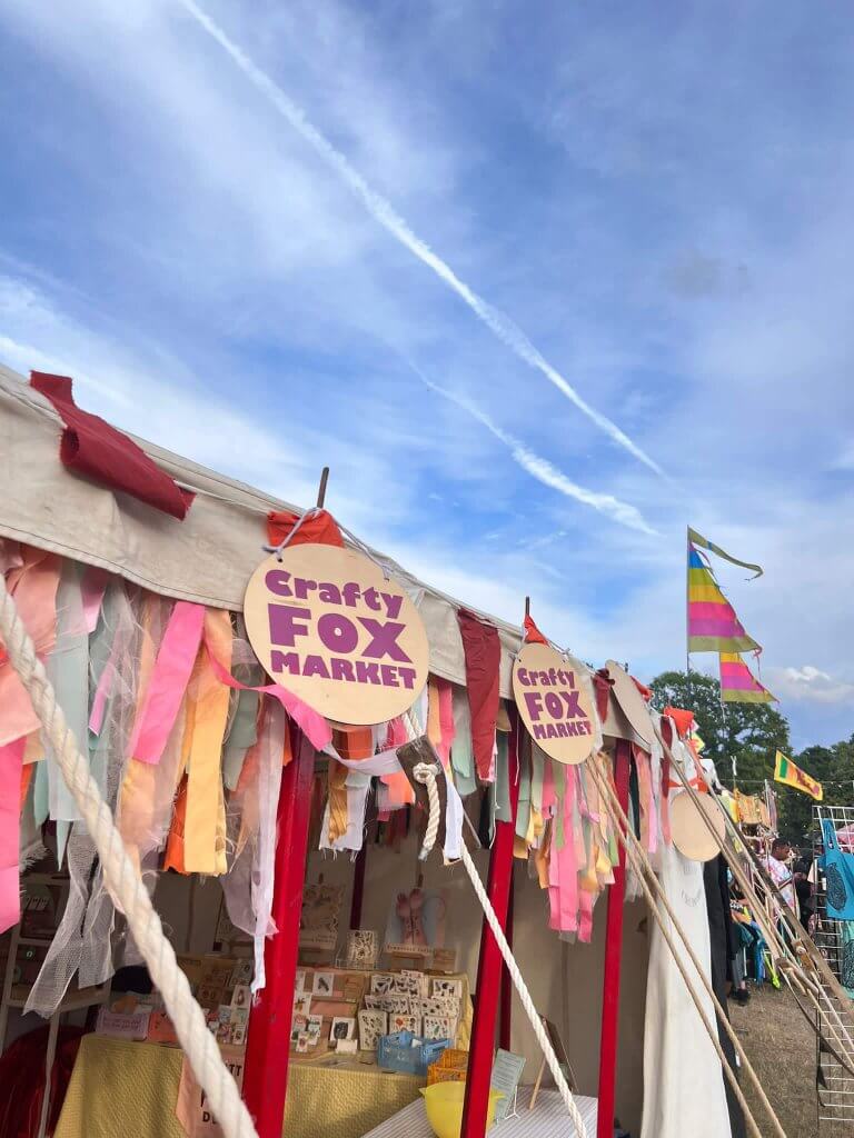 The open front of a large canvas marquee with bright red posts is adorned with pastel coloured bunting made of strips of fabric and round wooden signs printed in red with the words 'crafty fox market'. The sky above the tent is bright blue with some fluffy white clouds