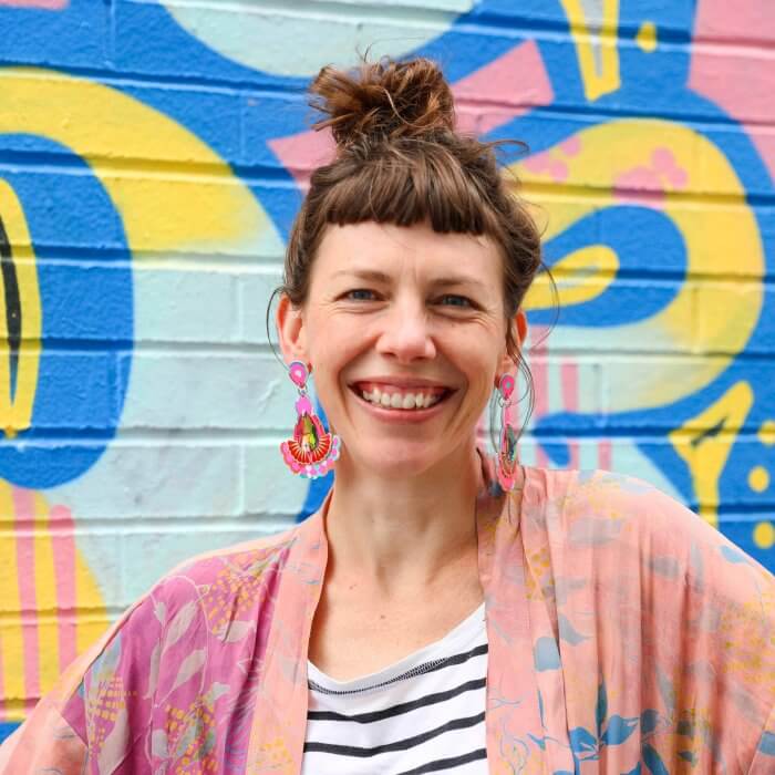 A woman with brown hair tied in a messy top knot, a short fringe and large bright pink statement earrings is smiling at the camera. Behind her is a colourful blue, pink and yellow graffiti covered wall