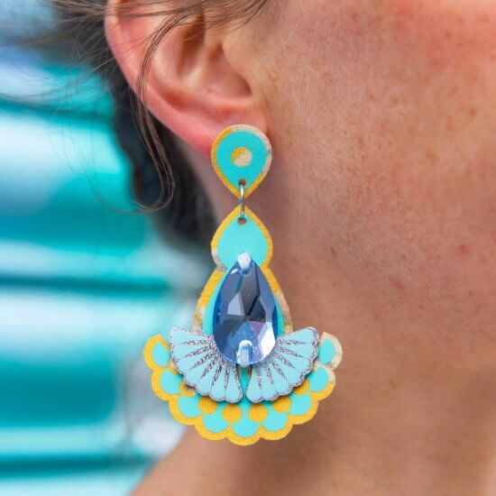 A close up of a woman's ear and neck, focusing on the blue, turquoise and yellow laser cut earrings she is wearing