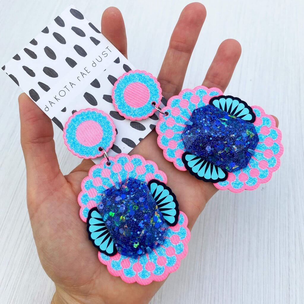 A pair of neon pink and bubblegum blue glittery giant jewel earrings are held in an open hand against an off white background