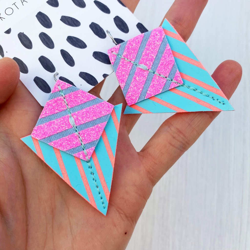 A pair of pastel pink, turquoise, peach and blue stripey geometric earrings held in a woman's open palm