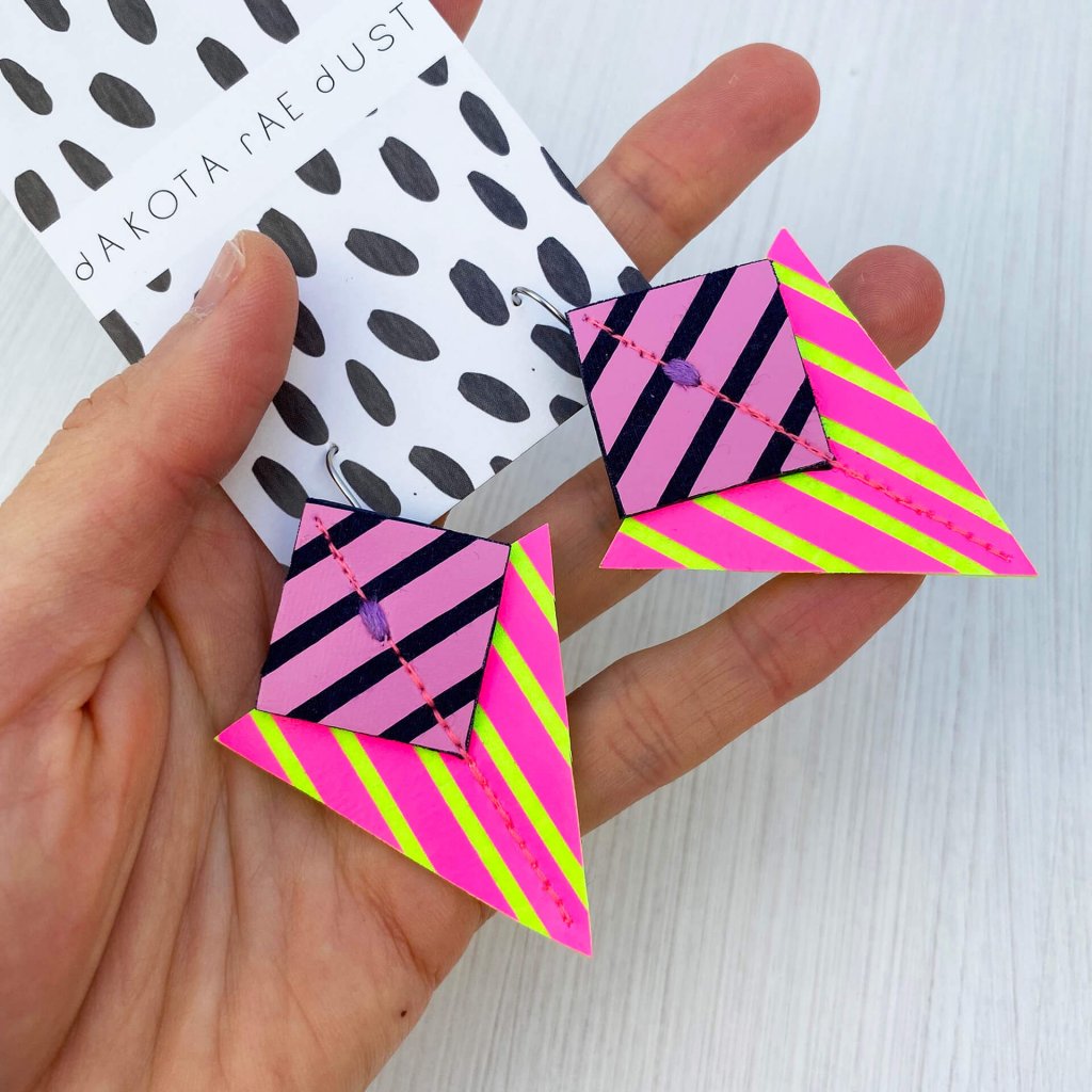 A pair of neon pink, purple and fluorescent yellow stripey geometric earrings held in a woman's open palm