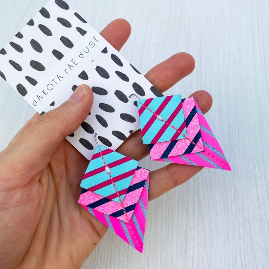 A pair of neon pink, blue and peacock green stripey geometric earrings held in a woman's open palm