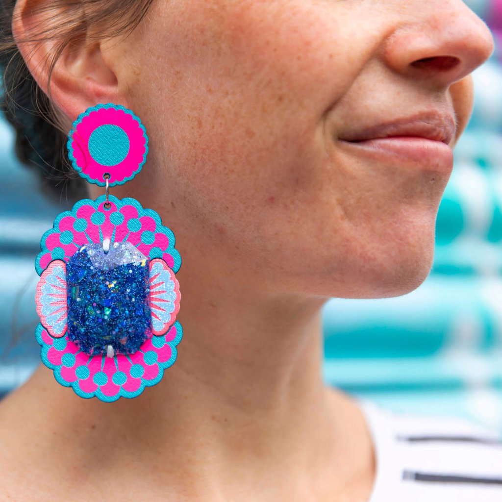 A close up of a woman's neck and ear, focusing on her colourful fluorescent pink and turquoise blue oversize jewel earrings.