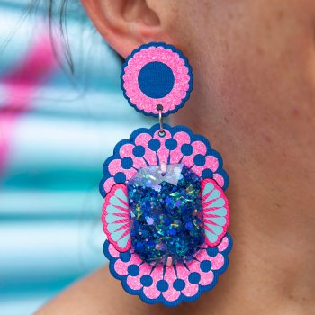 A close up of a woman's neck and lower ear focusing on her oversize dangly earrings in cobalt blue and glittery bubblegum pink, adorned with giant sapphire blue gems